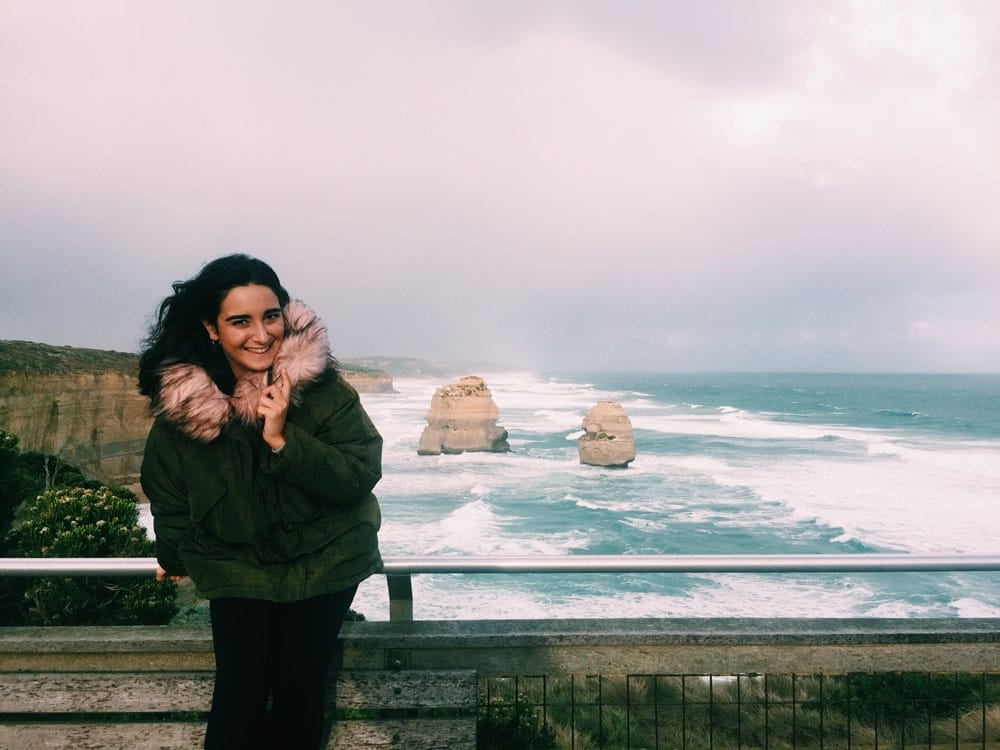 Judit en Melbourne chica morena con un abrigo verde sonriendo a cámara y detrás los apóstoles de Melbourne